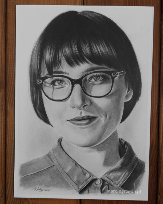 Portret wykonany ołówkiem. Portret przedstawia kobietę z ciemnymi, krótkimi włosami w okularach. Rysunek wykonany ba bloku Happy Color Rysunek.