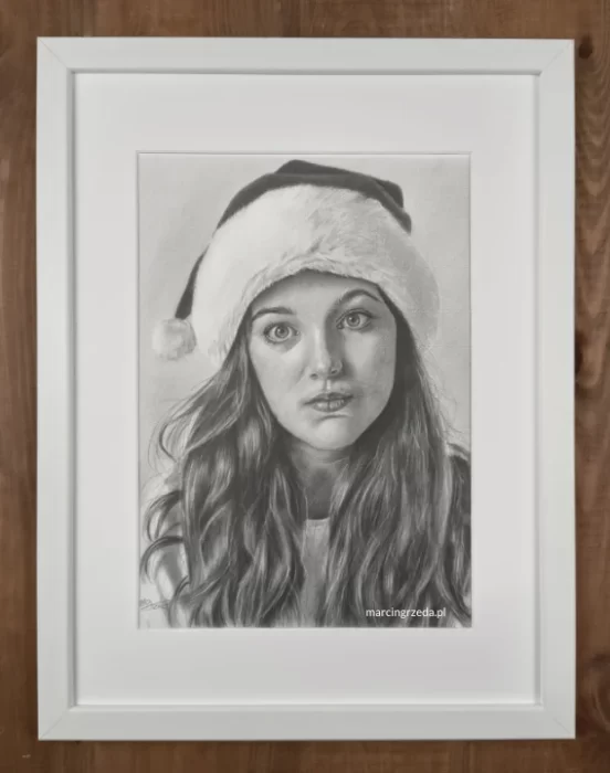 Portret pięknej dziewczyny wykonany ołówkiem oprawiony w białą ramkę z białym passe-partout. Dziewczyna ma na głowie czapkę Mikołaja.