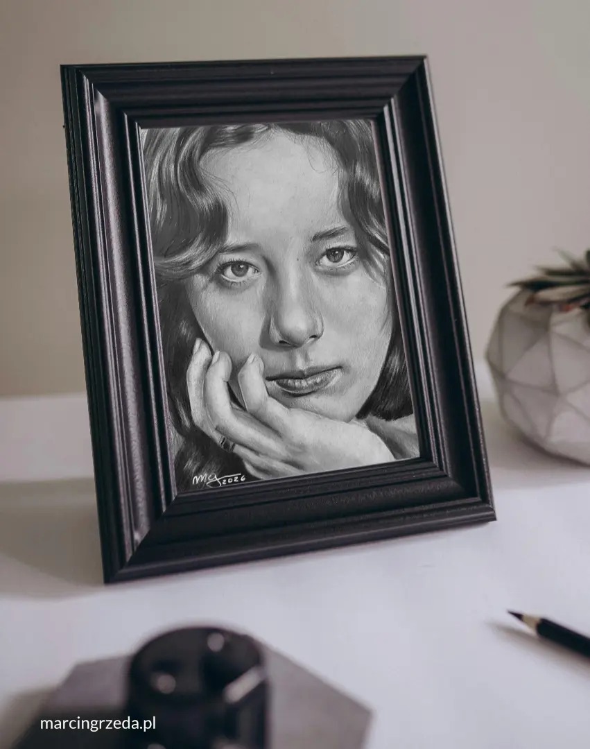 Portret wykonany ołówkiem ze zdjęcia umieszczony w ciemnej ramce. Portret stoi na stole. Jest to przykład sentymentalnego prezentu dla osoby, która już wszystko ma.